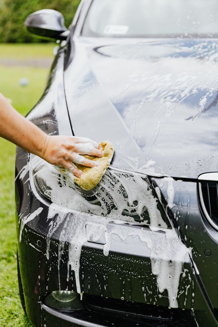 our-mission Person washing a black car's headlight with a sponge and soap outdoors.